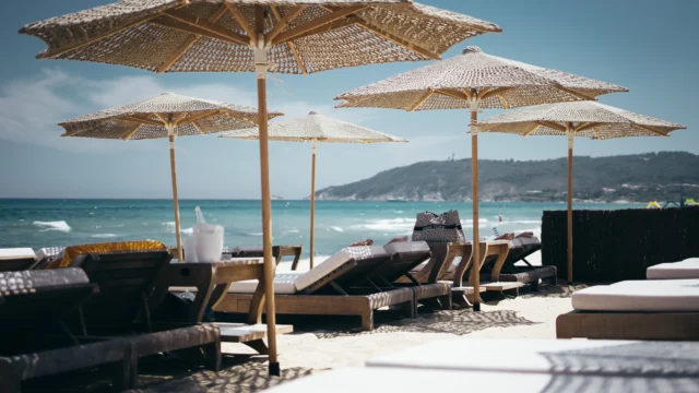 Exclusive beach club with woven rattan parasols and wooden sun loungers on white sand overlooking a turquoise sea with a green hillside in the background