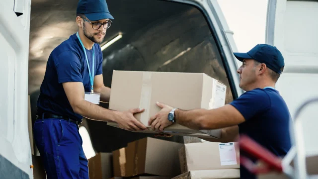 Two professional movers in blue uniforms unloading cardboard boxes from a delivery van
